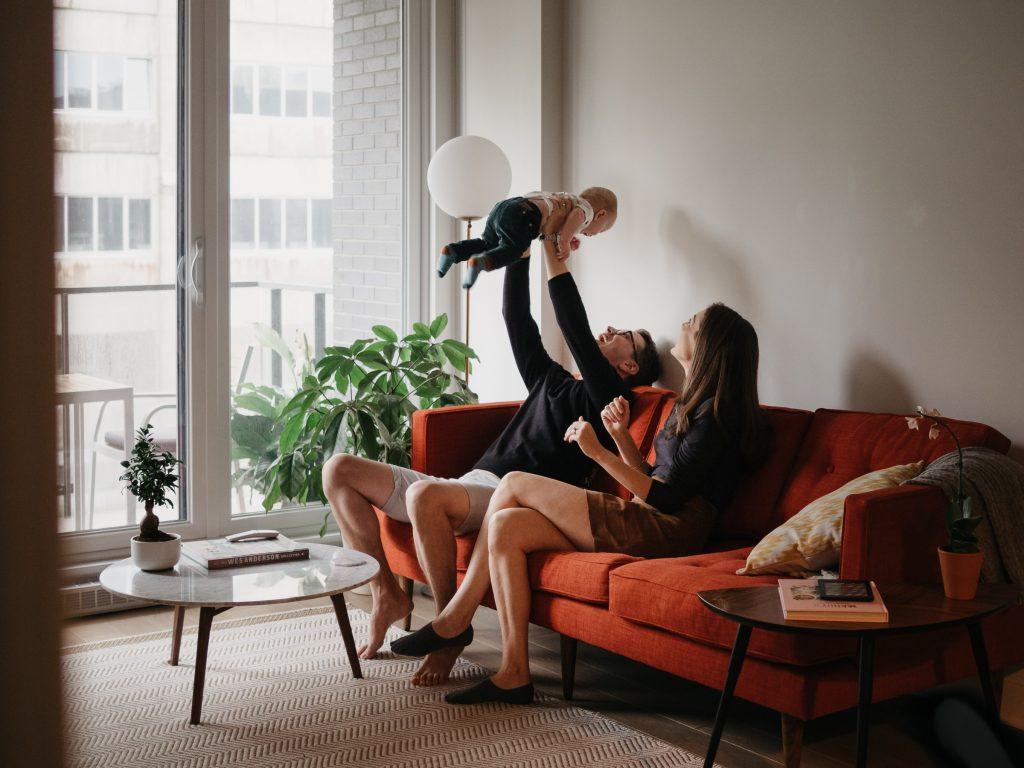 belle scène de complicité de parents assis sur le canapé pendant que le papa joue avec son nouveau-né lors d'une séance photo lifestyle à domicile