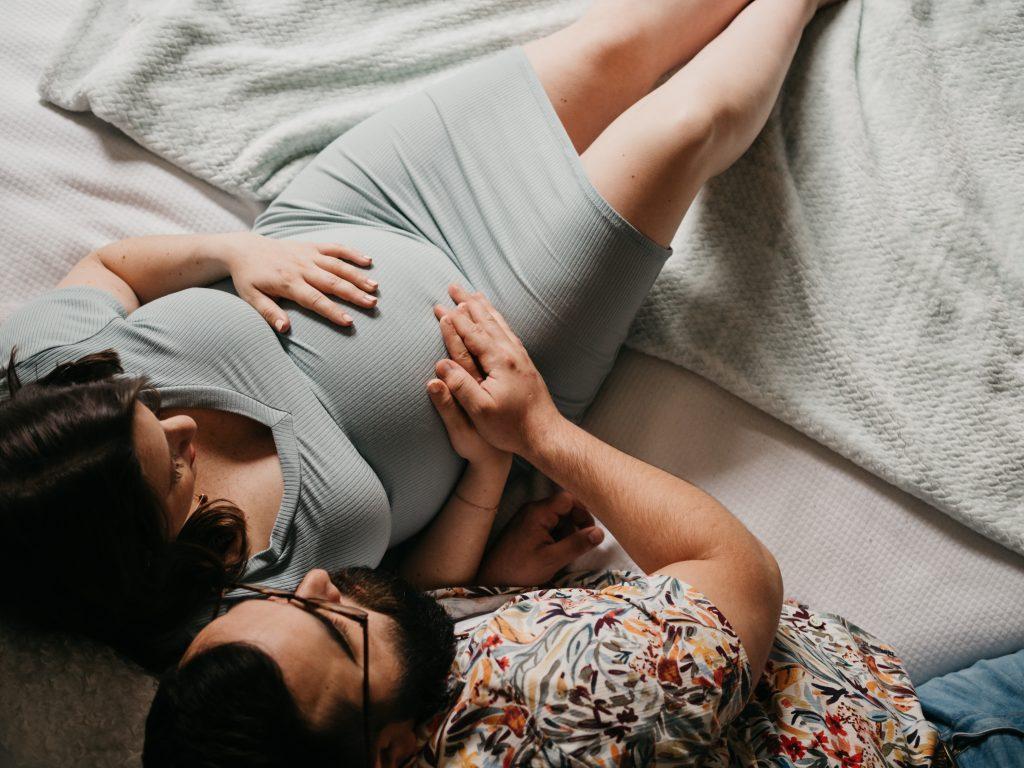 Couple allongé sur un lit se tenant le ventre lors d’une séance photo grossesse à domicile à Marseille