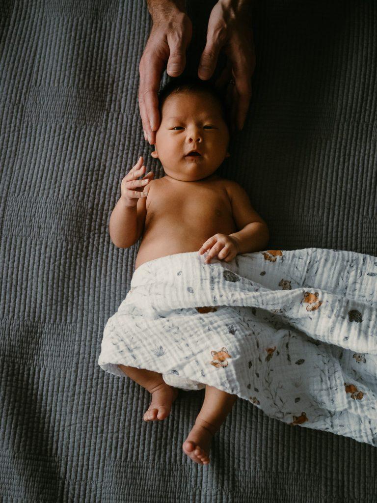 portrait de bébé nu sur un lit avec une couverture et les mains de son papa autour de la tête lors d'une session photo nouveau-né à domicile