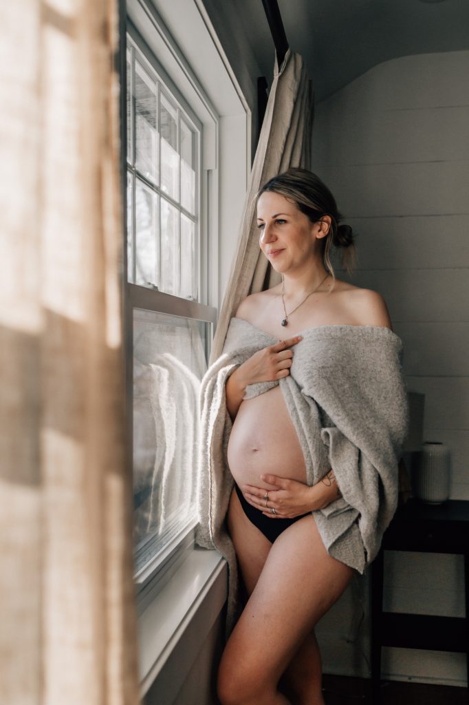 future maman en sous-vêtements caressant son ventre nu de femme enceinte devant une fenêtre lors d'une séance photo de grossesse boudoir à domicile à Marseille