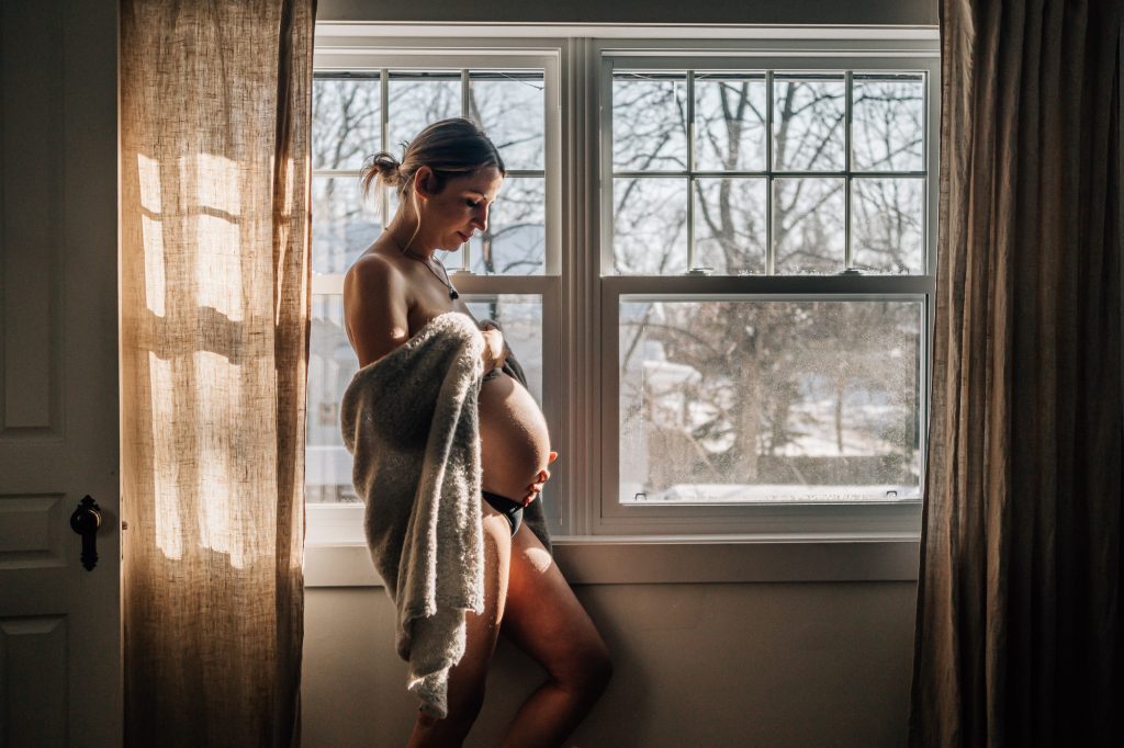 future maman en sous-vêtements caressant son ventre nu de femme enceinte devant une fenêtre lors d'une séance photo de grossesse boudoir à domicile à Marseille