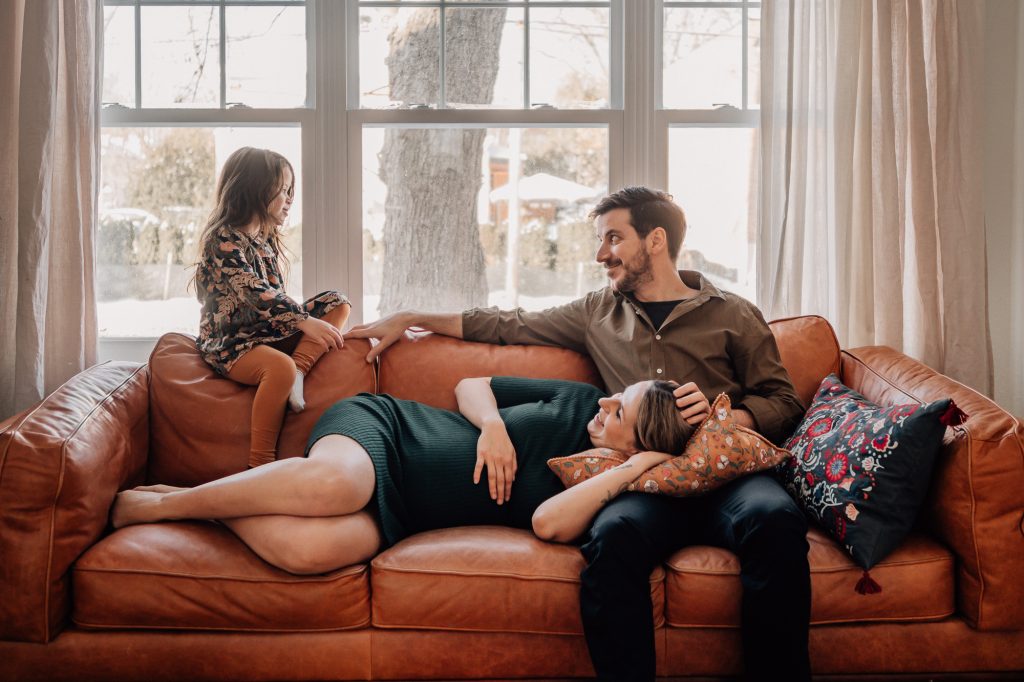 maman enceinte allongée sur son canapé et discute avec sa petite fille et son mari lors d'une séance photo lifestyle à domicile