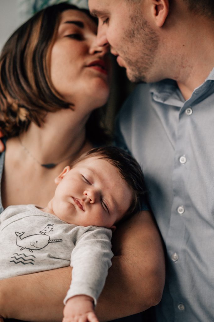 portrait de famille des parents qui s'embrassent en portant bébé qui dort dans les bras lors d'une séance photo nouveau-né à domicile à Marseille