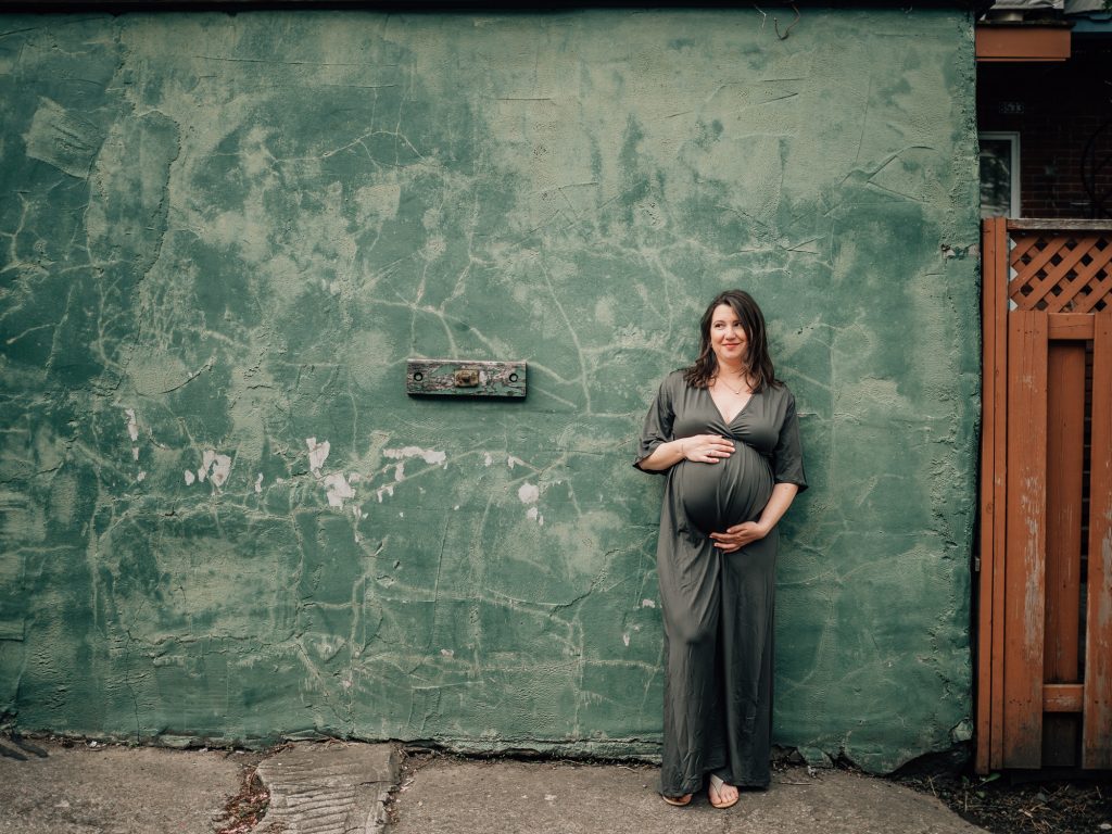 future maman adossée à un mur vert caressant son ventre rond lors d'une séance photo femme enceinte lifestyle en extérieur à marseille