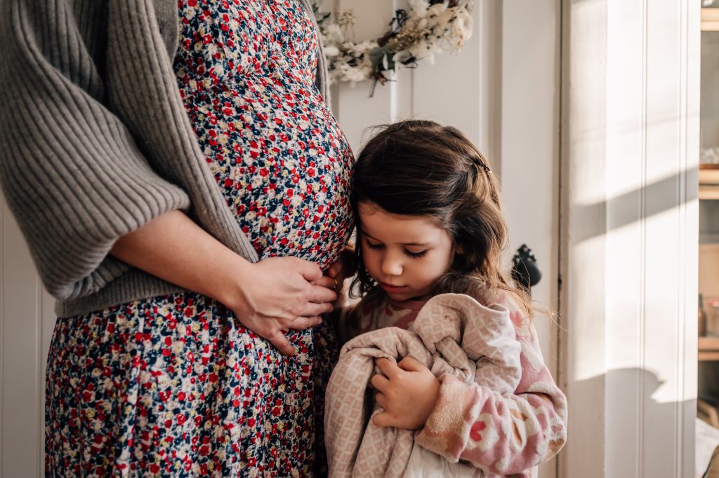 petite fille appuyant sa tête sur le ventre rond de sa maman enceinte qui porte une belle robe fleurie lors d'un shooting photo lifestyle de grossesse à domicile à marseille