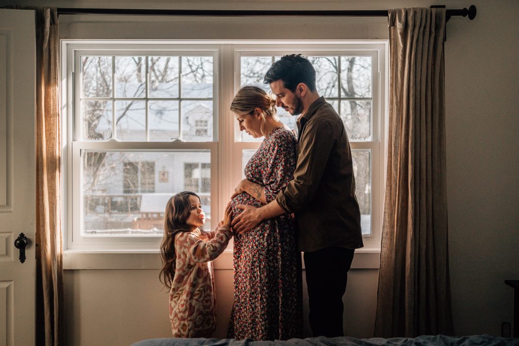 papa et petite fille touchant le ventre rond de la maman enceinte devant la fenêtre sous une belle lumière d'hiver lors d'un shooting photo grossesse à domicile à Marseille