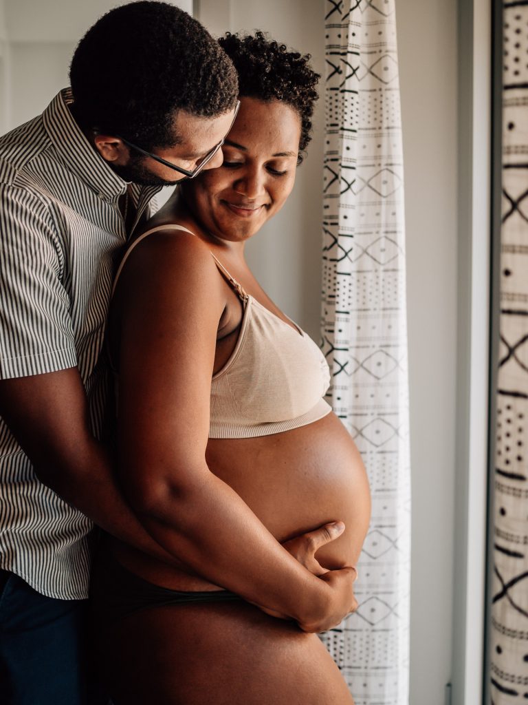 image extraite du portfolio de séance photo grossesse à domicile : future papa entourant le ventre rond de sa femme enceinte posant en sous-vêtements
