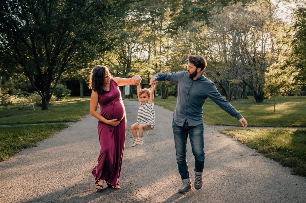 Illustration d'un couple de parents, dont la femme est enceinte, en train de faire sauter leur petite fille dans un parc à Marseille. Moment de complicité familiale et de jeu en plein air lors d'une séance grossesse lifestyle.