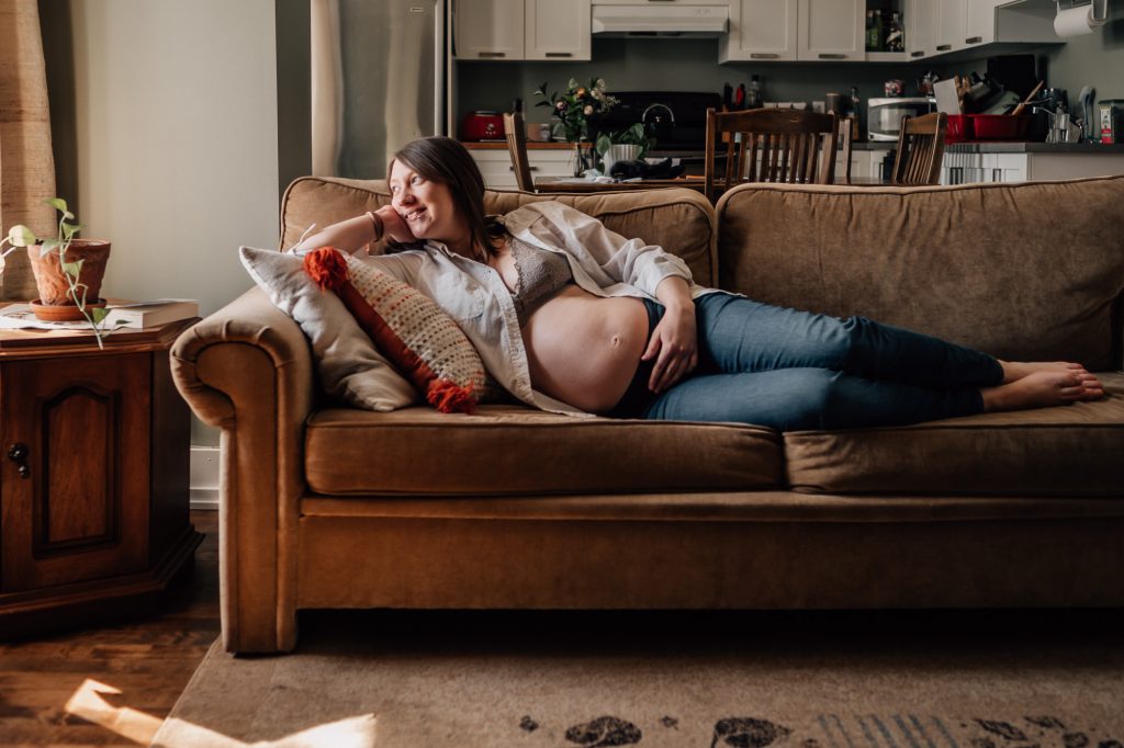 future maman dévoilant son ventre allongé sur son canapé et regarde par la fenêtre lors d'un shooting photo grossesse d'hiver à domicile à Marseille