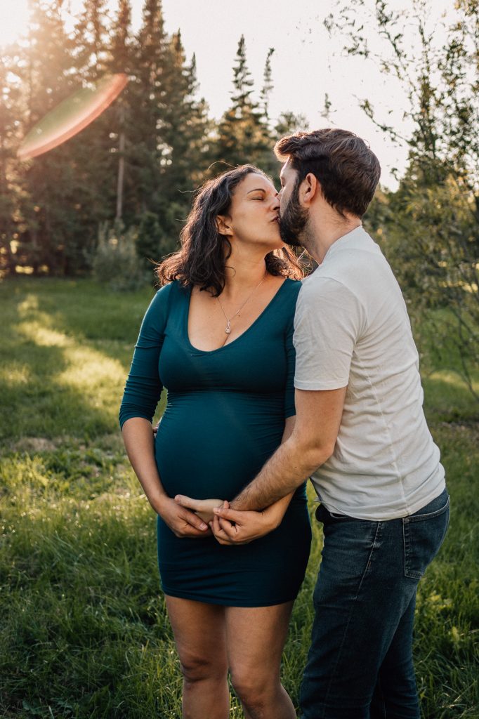 couple d'amoureux s'embrasse pendant que le papa pose sa main sur le ventre rond de la future maman dans un parc lors d'une séance photo grossesse lifestyle