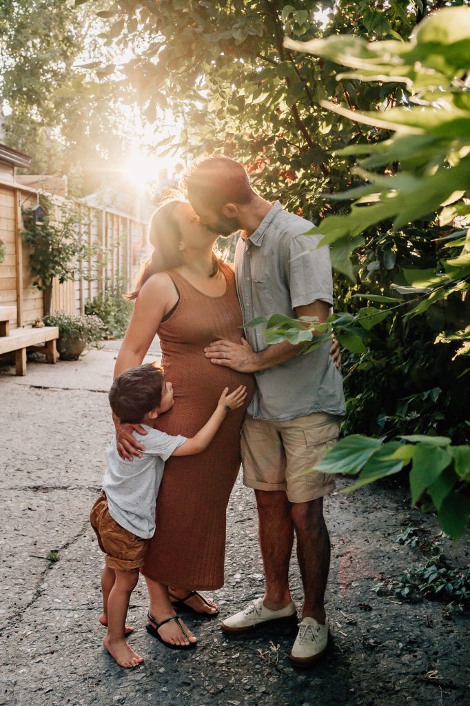 incroyable lumière de fin de journée sur une famille : le papa et l'enfant posent leur main sur le ventre de la maman enceinte pendant une mini séance de grossesse en extérieur