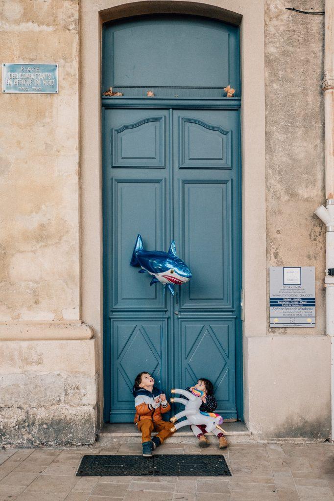 enfants jouant avec un ballon en forme de requin devant une porte bleue d'Aix-en-Provence, ville près de Marseille pendant un shooting photo en famille