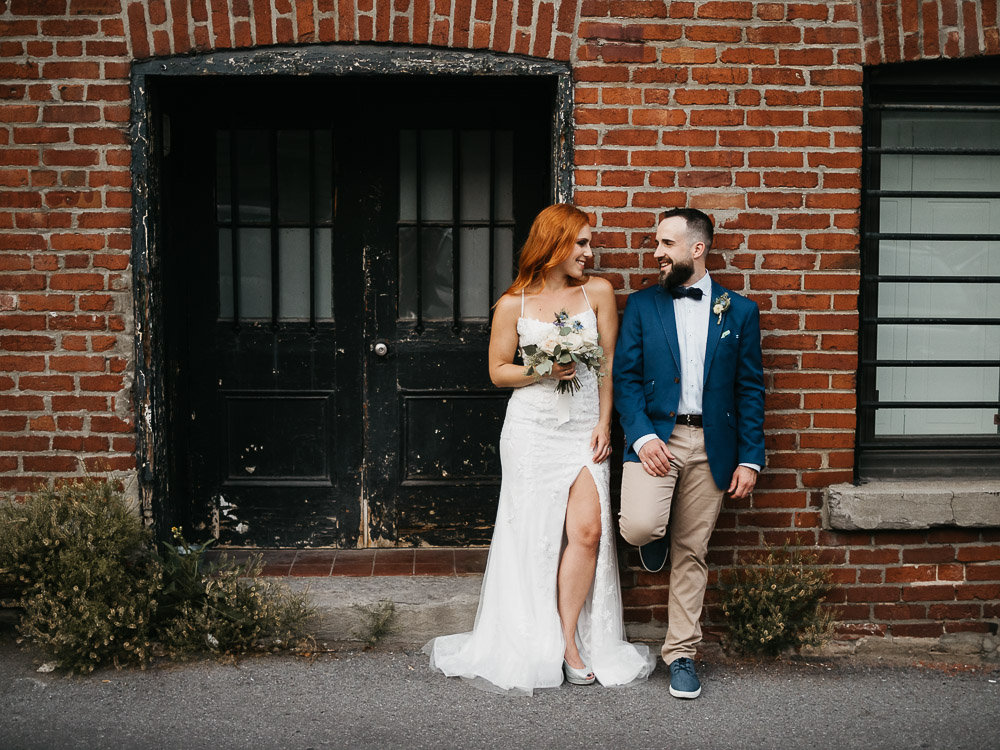 deux jeunes mariés heureux et souriants font un shooting photo après un mariage intime et engagement en ville devant un mur de briques rouges