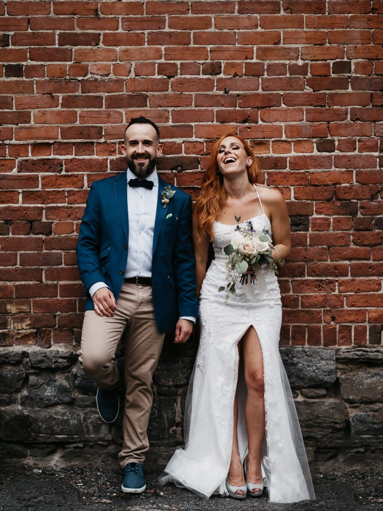 deux jeunes mariés heureux et souriants font un shooting photo après un mariage intime et engagement en ville devant un mur de briques rouges