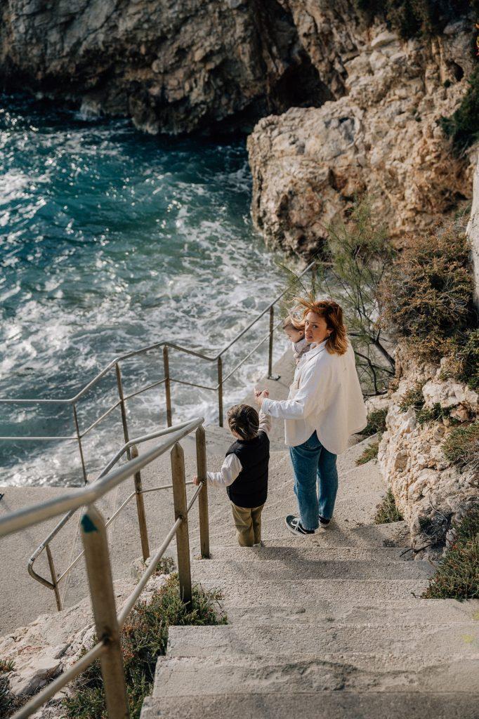 Maman qui descend les escaliers d'une crique donnant sur la mer avec son petit garçon lors d'une séance photo en famille à Malmousque à Marseille