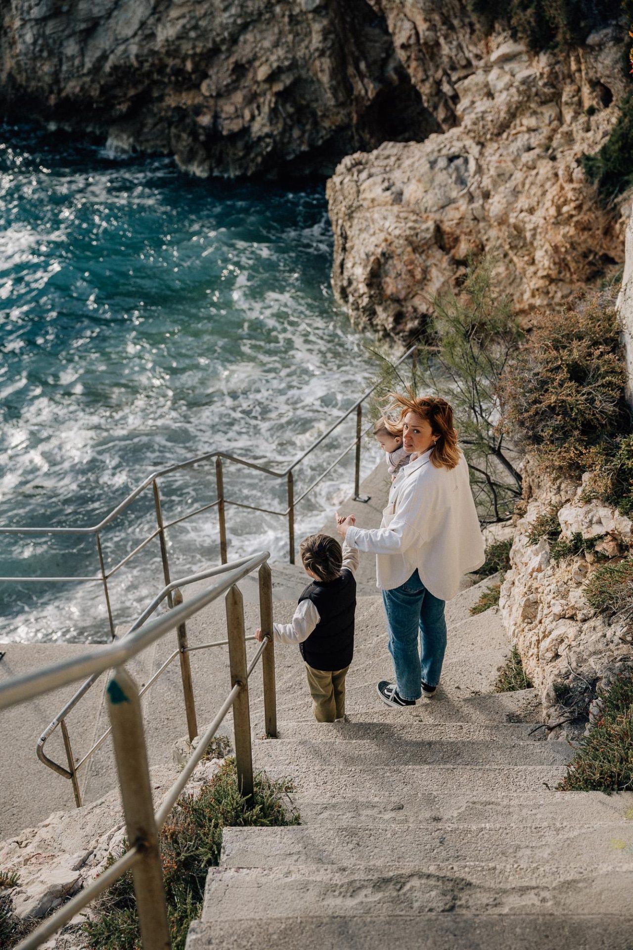 Maman qui descend les escaliers d'une crique donnant sur la mer avec son petit garçon lors d'une séance photo en famille à Malmousque à Marseille
