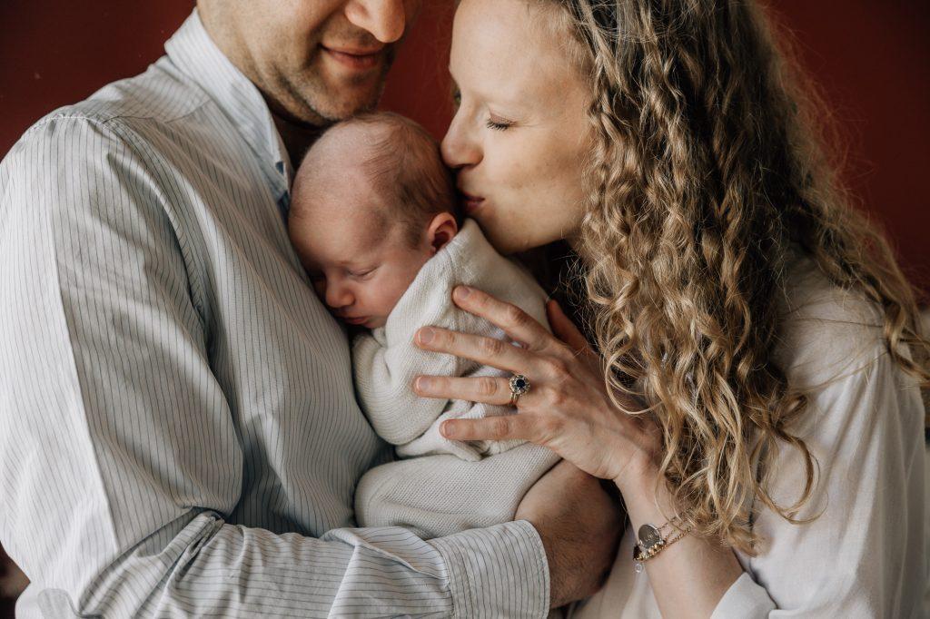 Maman embrasse son bébé dans les bras de son papa lors d'un shooting photo nouveau-né à domicile à Marseille 13007