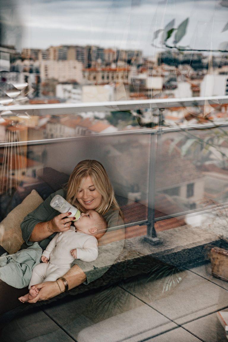 maman donne le biberon à son bébé lors d'une séance photo bébé à domicile, avec le reflet de Marseille dans la fenêtre