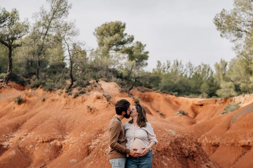 plan large d'un couple s'embrassant et caresse le ventre de la future maman lors d'un shooting grossesse à couple lors d'une séance photo de grossesse aux terres rouges à la sainte victoire près d'Aix-en-Provence