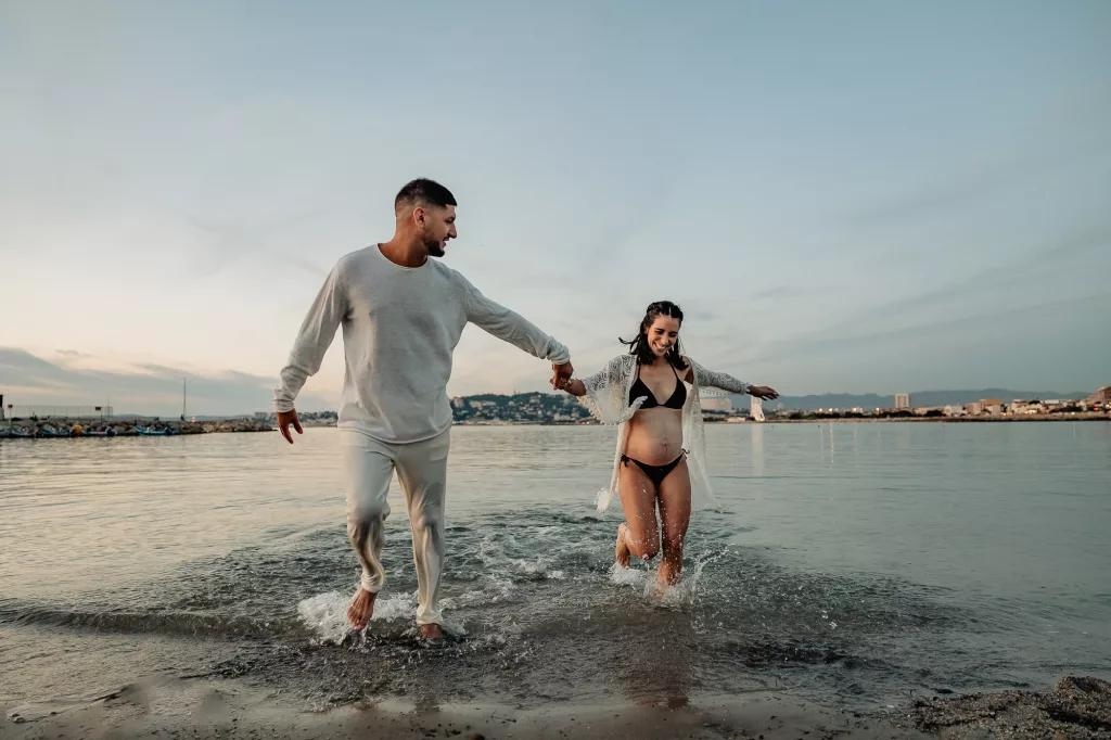 future maman court dans l'eau avec son mari à la plage de la Pointe Rouge lors d'un shooting photo grossesse à Marseille