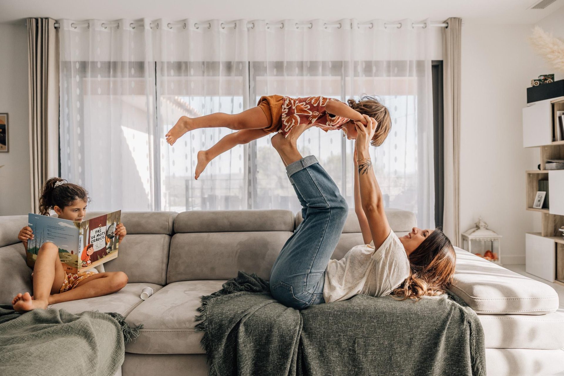 Petite fille qui lit un livre à côté de sa maman qui joue avec son petit frère sur le canapé lors d'une séance photo amusante lifestyle de famille à Marseille