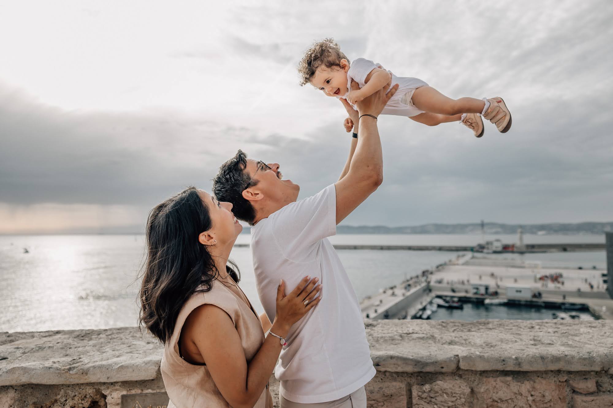 papa et maman lancent dans les airs leur petite fille pendant une séance photo lifestyle de famille au Mucem, vue sur la mer à Marseille