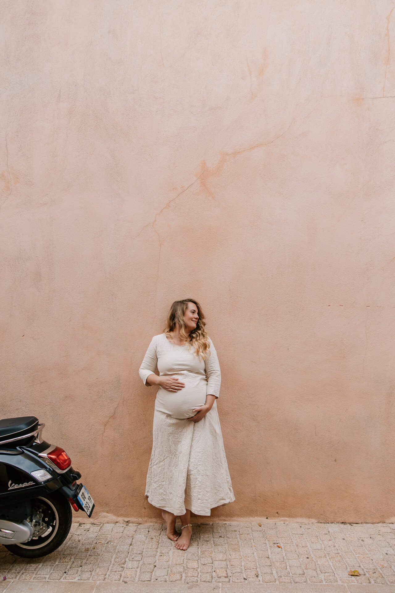 portrait de femme enceinte avec une longue robe beige devant un mur rose pastel et à côté d'un scooter au centre ville de Cassis