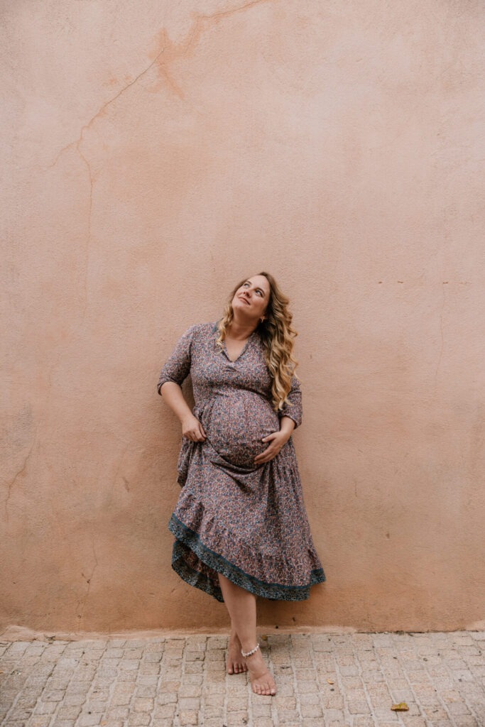 Femme enceinte avec une robe fleurie appuyée contre un mur ocre lors d’une séance photo à Cassis