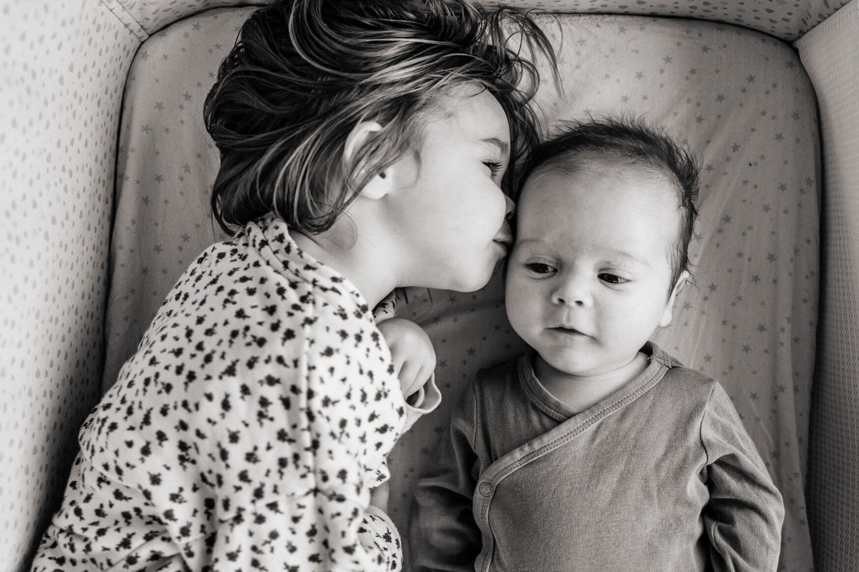 Grande soeur fait un bisou à son petit frère allongés côte à côte lors d’une séance photo bébé à domicile à La Ciotat