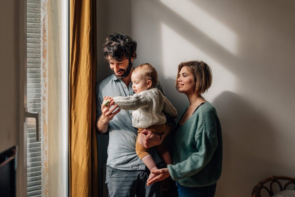Parents avec leur bébé d'un an près d’une fenêtre lors d’une séance photo bébé à domicile à Marseille 13006