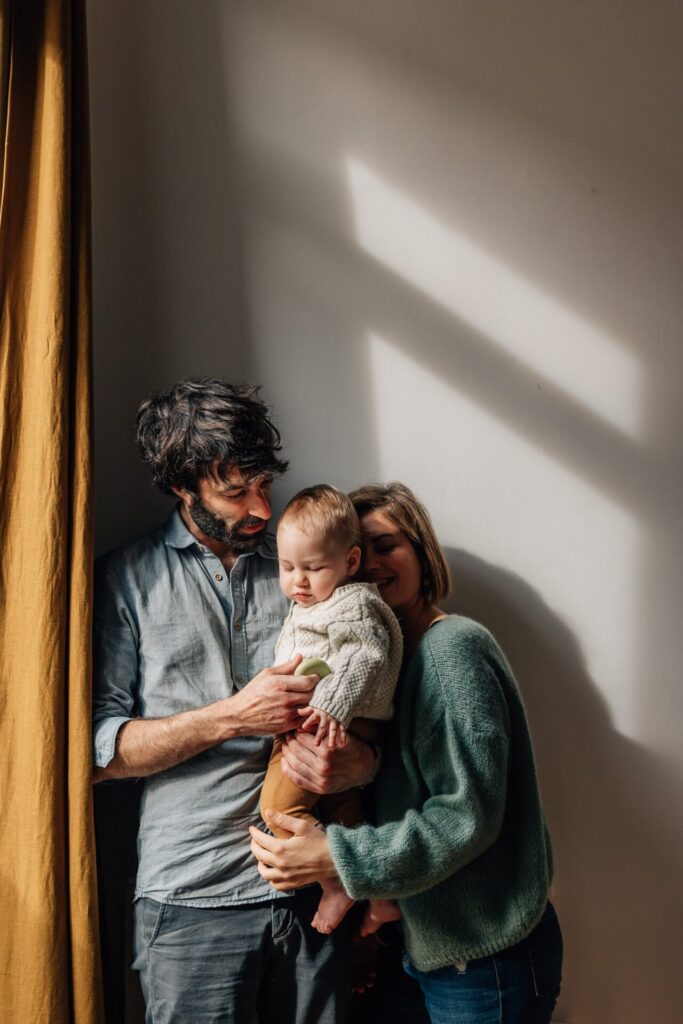 Parents et bébé serrés près d’une fenêtre, lumière du matin, séance photo à domicile à Marseille