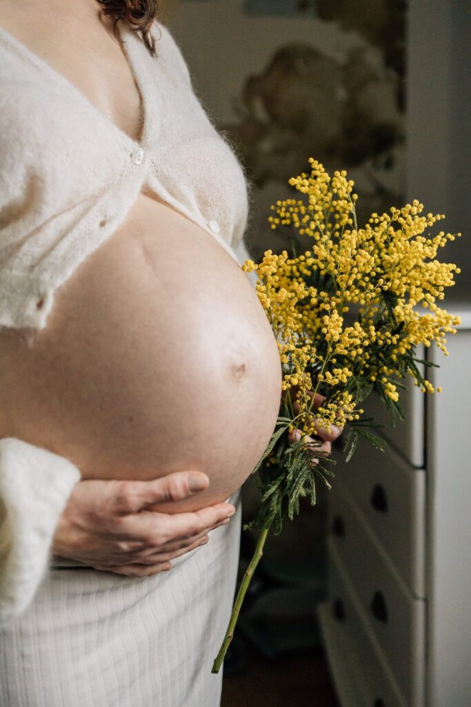 Gros plan sur le ventre d'une femme enceinte qui tient un bouquet de mimosa dans sa main lors d'une séance photo grossesse à domicile - Inspiration shooting créatif fleuri