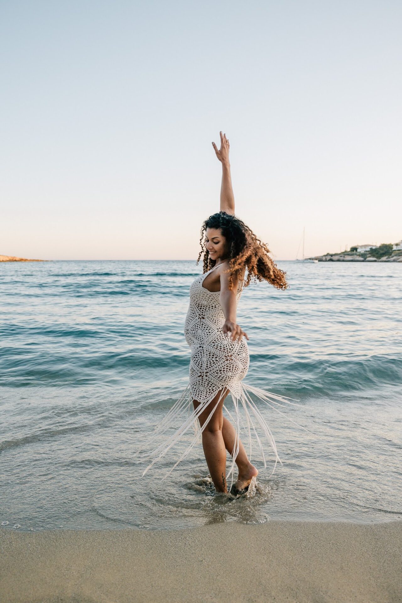Femme enceinte en robe claire dansant dans l’eau lors d’une séance photo grossesse à la plage au coucher du soleil