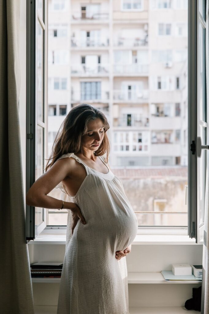 Femme enceinte près d’une fenêtre avec vue sur les immeubles de Marseille lors d’une séance photo grossesse à domicile