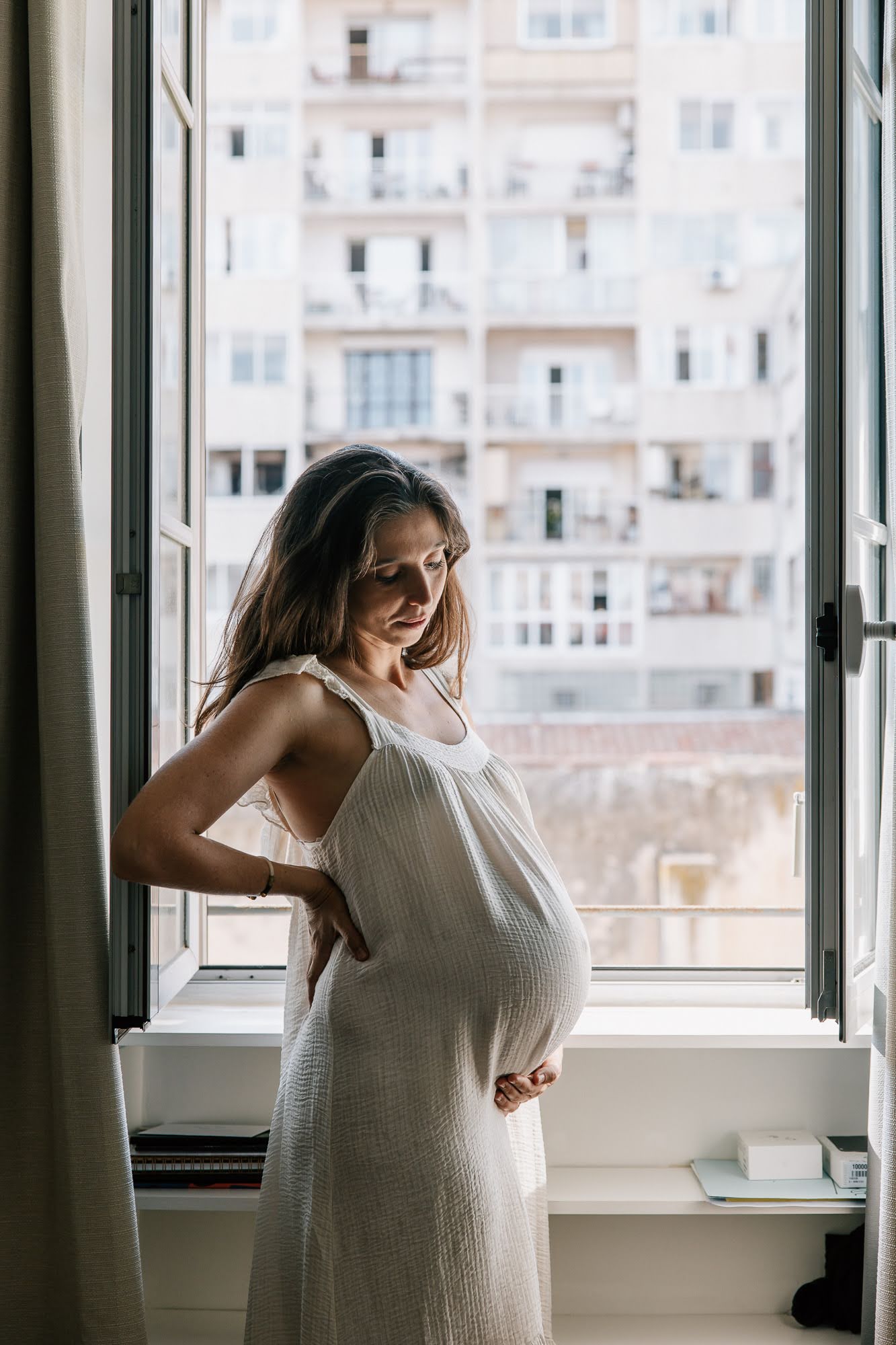 Femme enceinte près d’une fenêtre avec vue sur les immeubles de Marseille lors d’une séance photo grossesse à domicile