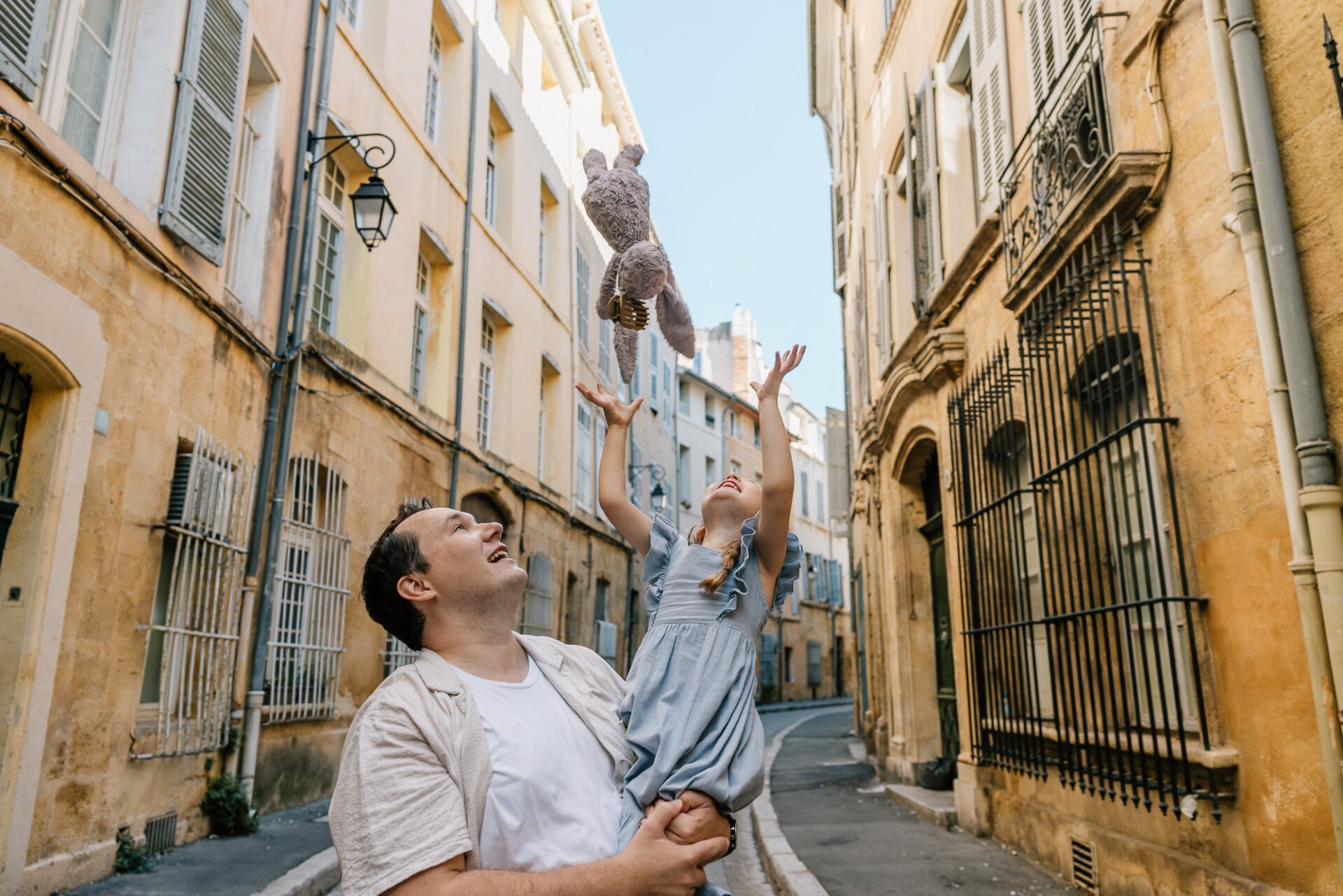 père portant sa fille dans une rue pavée à aix en provence ours en peluche lancé en l’air