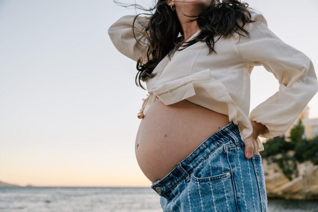 détail du ventre d’une femme enceinte en jean face à la mer lors d’une séance photo grossesse
