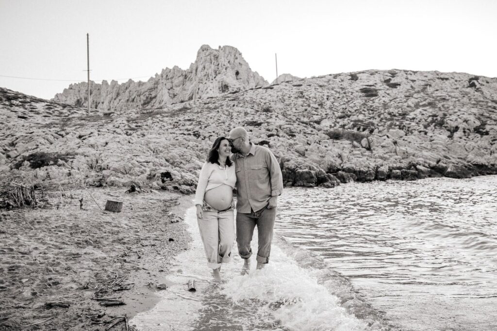 Couple marchant au bord de l'eau pendant un shooting photo grossesse à la plage à Marseille 13008