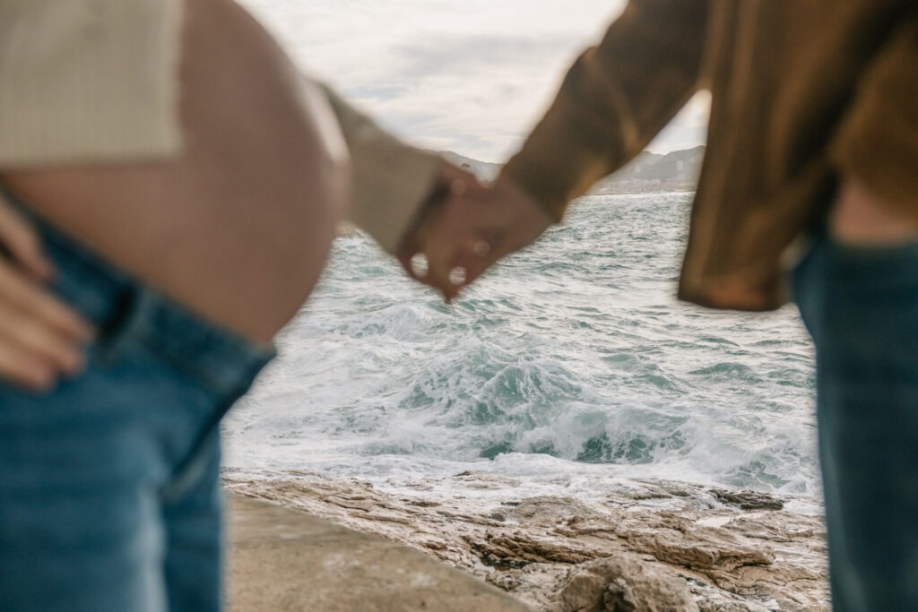 Détail des mains et ventre en séance grossesse à Marseille, flou artistique et mer en arrière-plan