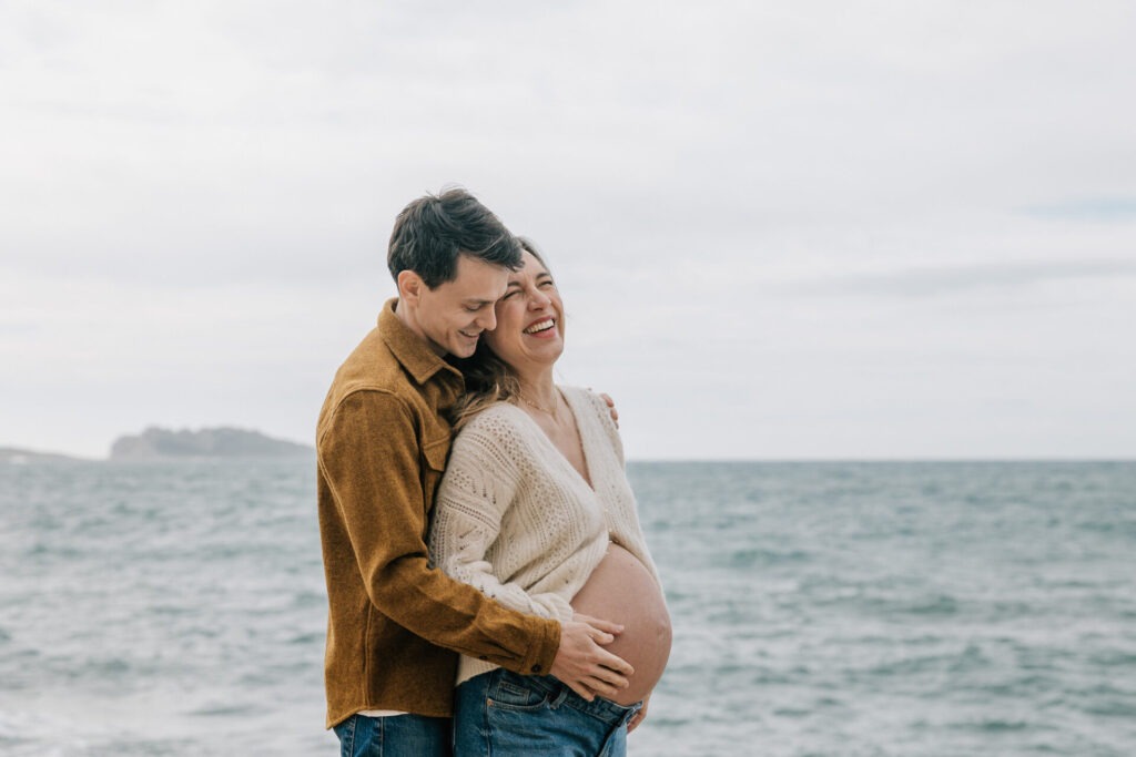 Couple heureux enlacé en séance grossesse à Marseille, lumière naturelle et mer agitée en arrière-plan