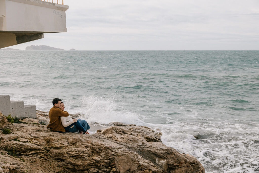 Couple en séance grossesse à Marseille assis sur les rochers en bord de mer, regardant les vagues