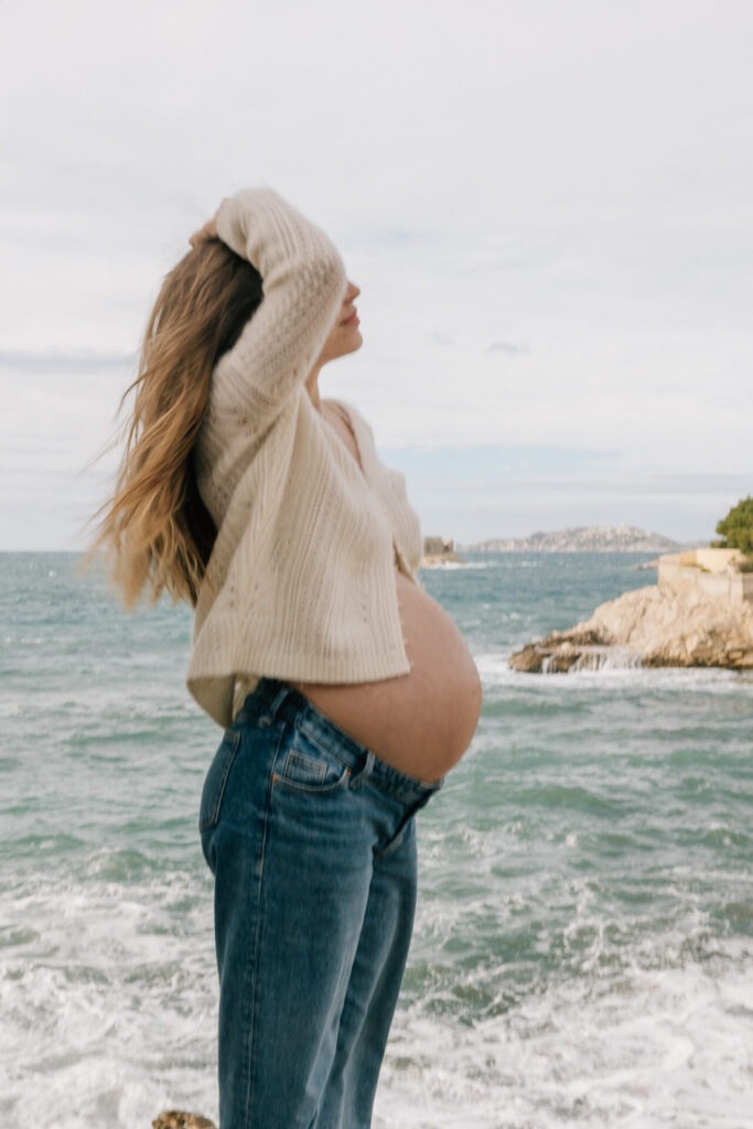 Portrait de grossesse en profil à Malmousque à Marseille, vent dans les cheveux face à la mer agitée, ambiance naturelle et lumière diffuse sous ciel nuageux